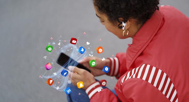 A person on a phone, which is surrounded by a whirlwind of social media channel icons to represent the different platforms a company can outsource the marketing for to a social media marketing agency.